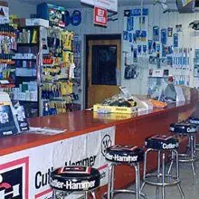 An older colour photograph of the Arrow sales counter, with Cutler-Hammer branded stools and wall-mounted product pegboards.