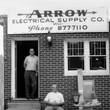 A black and white photograph of the original Arrow Electrical Supply Co. storefront, with a hand-painted sign and two men standing in the doorway.